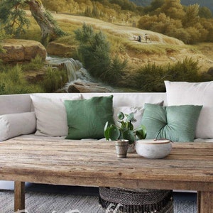 Op de afbeelding: Een rustieke houten salontafel met een landschapsschildering op de achtergrond. De tafel is versierd met witte en groene kussens, een kleine plant en een decoratieve kom. Onder de tafel staat een geweven mand.