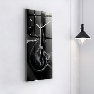 Pode incluir: Um relógio com um design abstrato em preto e branco, com ponteiros prateados e fundo branco. O relógio é emoldurado em uma moldura de vidro preto.