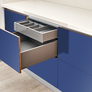 May include: Contemporary kitchen cabinets in a deep blue hue. The open drawer shows a grey cutlery organiser. The cabinet doors have a wood grain edge. The countertop is white, and the floor is light-coloured wood.