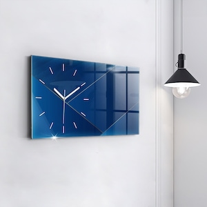 Puede incluir: Un reloj de pared moderno con un diseño geométrico. La esfera del reloj es azul oscuro con detalles dorados y manecillas blancas. El reloj rectangular está montado en una pared blanca.