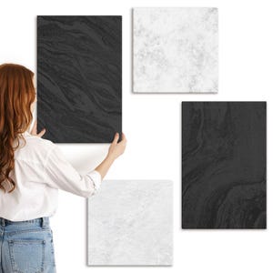 May include: A person holding a black rectangular wall art with a marbled design. Three other square wall art pieces are visible, one black, one white with a marbled design, and one white with a speckled design. The wall art pieces are displayed against a white wall.
