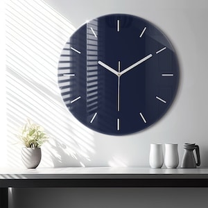 May include: A large, round, navy blue wall clock with white hour and minute hands and white markings for the hours. The clock is hanging on a white wall.