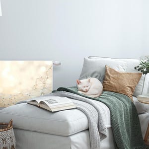 Puede incluir: Un gato blanco acurrucado en una chaise longue blanca con una manta de tiro verde. Un libro está en la chaise longue y una mesa auxiliar de madera con un jarrón de flores blancas está en el fondo.