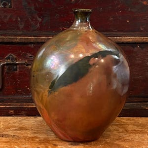 May include: A handcrafted, iridescent glass bottle with a rounded body and a narrow neck. The bottle features a blend of brown, copper, and green hues, with a dark, organic-shaped accent. The surface reflects light, creating a shimmering effect.