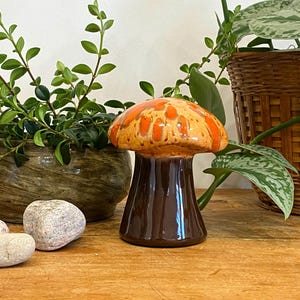 May include: A ceramic mushroom figurine with a brown stem and a cap in shades of orange and tan. The mushroom is displayed on a wooden surface, with green plants and rocks in the background. The figurine is a decorative item.