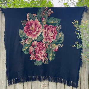 May include: A black blanket with a large floral design featuring pink and red roses, green leaves, and yellow accents. The blanket has a fringe trim along the edges. The blanket is hanging on a wooden fence.