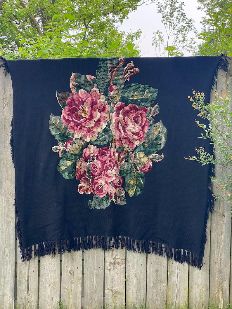 May include: A black blanket with a large floral design featuring pink and red roses, green leaves, and yellow accents. The blanket has a fringe trim along the edges. The blanket is hanging on a wooden fence.