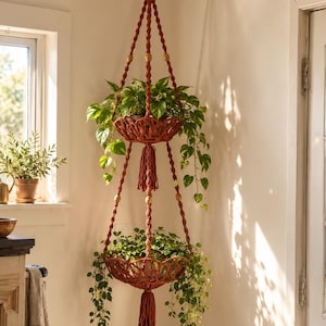 May include: A two-tier macrame plant hanger in a deep rust colour featuring knotted cord construction, wooden beads, and two hanging baskets. Each tier holds a green leafy plant with trailing vines, and the structure ends in a long tassel.