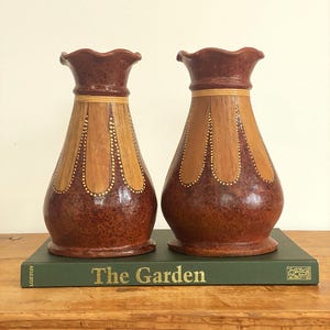 May include: A pair of brown ceramic vases with a wood-like pattern and gold accents. The vases have a scalloped rim and are sitting on a green book titled "The Garden".