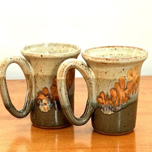 May include: Two handmade ceramic mugs with large handles. The mugs have a speckled beige and brown glaze with orange and brown floral designs. The mugs are on a wooden surface.