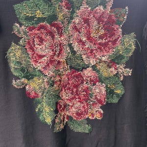 May include: Black textile with a large floral design. The flowers are primarily shades of red and pink, with green leaves and accents of yellow. The textile has a fringe along the bottom edge.