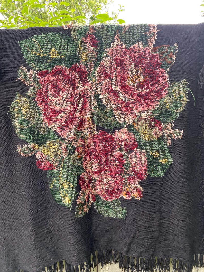 May include: Black textile with a large floral design. The flowers are primarily shades of red and pink, with green leaves and accents of yellow. The textile has a fringe along the bottom edge.
