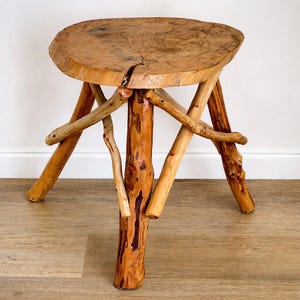 Może przedstawiać: Rustykalny drewniany stołek z okrągłym blatem i trzema nogami. Blat wykonany jest z jednego kawałka drewna, a nogi z gałęzi. Stołek ma około 46 cm wysokości.