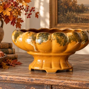 May include: A glazed ceramic bowl with a scalloped rim and a pedestal base. The bowl features a glossy mustard yellow finish with dark green sponged accents along the upper edge and a wide, rounded body shape supported by a sturdy, fluted base.