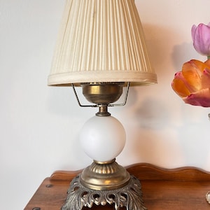 Peut inclure: Une lampe de table vintage avec un abat-jour plissé de couleur crème. La lampe présente une base en laiton avec un design orné, un globe blanc et un luminaire en laiton. La lampe est sur une surface en bois.