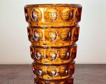 May include: Amber-colored glass vase with a geometric, textured design. The vase has a cylindrical shape with a series of stacked, hexagonal sections, each adorned with circular, embossed patterns. The vase is made of thick glass.