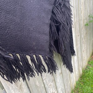 May include: A black woven shawl with long fringe detail. The shawl is draped over a wooden fence, showcasing the texture of the fabric and the fringe along the edges. The fringe adds a decorative element.