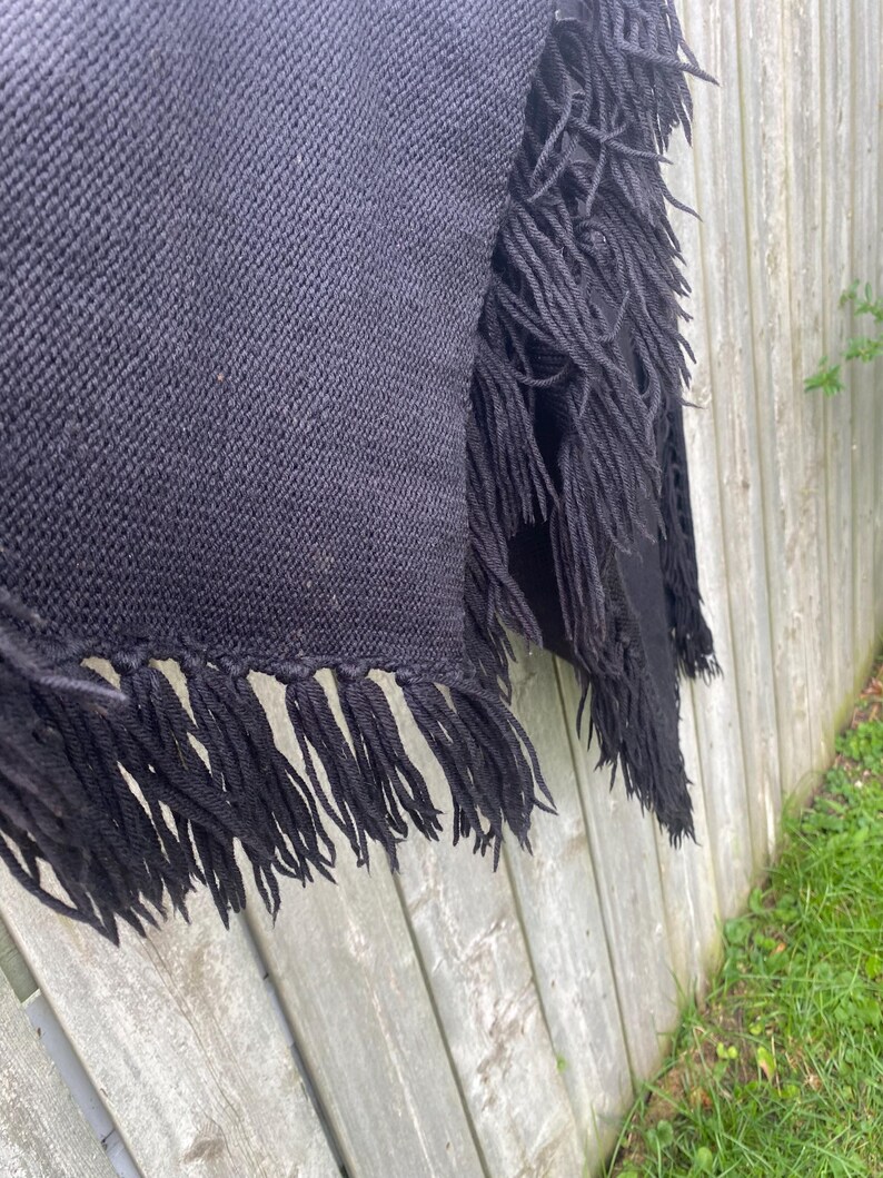 May include: A black woven shawl with long fringe detail. The shawl is draped over a wooden fence, showcasing the texture of the fabric and the fringe along the edges. The fringe adds a decorative element.