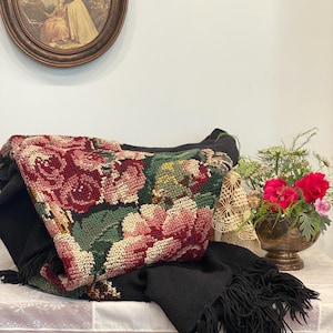 May include: A black knitted throw blanket with a floral tapestry design featuring red and pink roses, green leaves, and a black fringe. The blanket is draped over a table with a white lace tablecloth.
