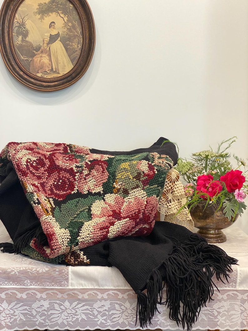 May include: A black knitted throw blanket with a floral tapestry design featuring red and pink roses, green leaves, and a black fringe. The blanket is draped over a table with a white lace tablecloth.