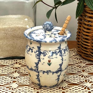 May include: A ceramic sugar bowl with a lid and a wooden spoon. The bowl is white with a blue plaid pattern and a small red and green floral design. The lid has a blue knob.