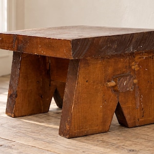 Könnte beinhalten: Ein niedriger, rechteckiger Holzhocker mit einer dicken, dunkelbraunen Sitzfläche und hellbraunen, angewinkelten Beinen. Der Hocker weist eine sichtbare Holzmaserung, ein verwittertes Finish und eine robuste Konstruktion mit Verbindungsdetails auf.