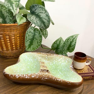 Puede incluir: Un plato de cerámica vintage con dos compartimentos, con interior verde claro con motas blancas y exterior marrón. Una taza pequeña marrón y blanca y una planta en una cesta tejida están en el fondo.
