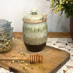 May include: A handmade ceramic honey pot with a lid and wooden honey dipper. The pot has a green and brown glaze. A wooden chopping board holds the honey dipper and a small amount of honey. Dried chamomile flowers are scattered around the board.