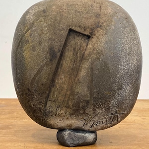 May include: An abstract sculpture with a rounded, oval form. The sculpture is predominantly brown with darker markings and a rectangular cut-out. It is mounted on a small, dark grey base. The artist's signature is visible.