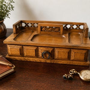 May include: A rectangular wooden desk organiser featuring a natural brown wood grain finish. The top surface includes three recessed compartments and a lattice-style back rail. A single drawer is centred on the front, accented with a horseshoe handle.