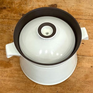 May include: A white ceramic casserole dish with a lid. The dish has a brown interior and rim, with a white lid featuring a brown and white circular design. The dish has two white handles and sits on a white base. The overall design is simple and functional.