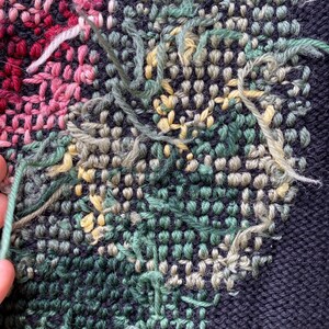 May include: Close-up of a textile with a floral design. The piece features a dark blue background with embroidered flowers in shades of pink, green, and yellow. Loose threads add texture to the handcrafted artwork.