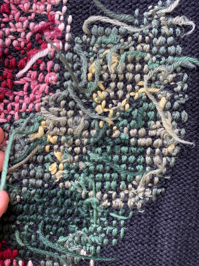 May include: Close-up of a textile with a floral design. The piece features a dark blue background with embroidered flowers in shades of pink, green, and yellow. Loose threads add texture to the handcrafted artwork.