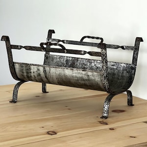 May include: An antique metal magazine rack with a distressed finish. The rack features ornate scrollwork on the legs and sides, with a handle on top. The interior is a light colour, contrasting with the darker metal frame. The rack is on a wooden surface.