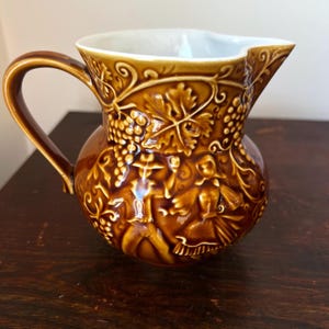 May include: A brown ceramic jug with a handle and spout. The jug features a relief design of grapes, leaves, and figures. The interior is white. The jug is on a dark wooden surface.