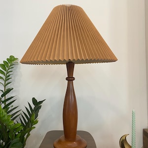 Op de afbeelding: Een vintage tafellamp met een geplooide beige lampenkap en een donkerbruine houten voet. De lamp heeft een klassiek, modern design uit het midden van de eeuw. De lamp staat op een houten tafel, met een groene plant en een kandelaar op de achtergrond.