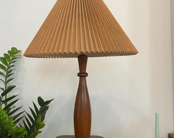 May include: A vintage table lamp with a pleated tan lampshade and a dark brown wooden base. The lamp has a classic, mid-century modern design. The lamp is on a wooden table, with a green plant and a candle holder in the background.