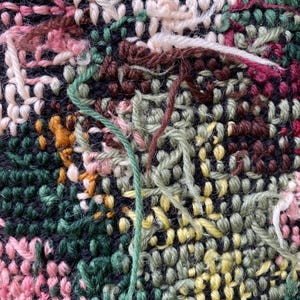 May include: Close-up of a textured textile with a complex weave. The fabric features a mix of colors, including pink, green, yellow, and brown, with visible yarn strands. The background is a dark navy blue.