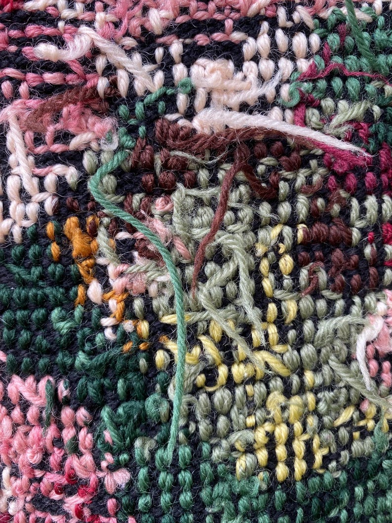 May include: Close-up of a textured textile with a complex weave. The fabric features a mix of colors, including pink, green, yellow, and brown, with visible yarn strands. The background is a dark navy blue.