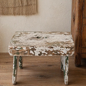 May include: A small, square wooden stool with a distressed, aged appearance. The stool has a white, peeling paint finish over a brown base, with hints of green. It has four short legs and a hole in the centre of the seat.