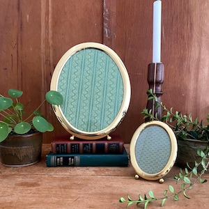 May include: Two oval gold-toned picture frames with green patterned inserts. One frame is larger and rests on a stack of books. A small frame sits on a wooden surface. A white candle sits in a dark wooden holder.