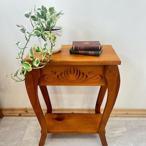 Peut inclure: Une table d'appoint en bois avec un motif décoratif sculpté sur le devant. La table a des pieds incurvés et une étagère inférieure. Une plante en pot avec des feuilles vertes et blanches et deux livres sont sur le dessus.
