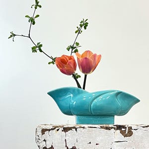 Könnte beinhalten: Eine türkisfarbene Keramikvase mit einem einzigartigen, fließenden Design, die zwei orangefarbene und rosa Tulpen und grüne Zweige enthält. Die Vase ist mit "MADE IN CALIFORNIA H-15" beschriftet und steht auf einer weißen, abgenutzten Oberfläche.