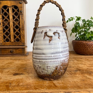 May include: A white ceramic vase with a brown, woven handle. The vase has a rustic, textured finish and is decorated with brown and white stripes.