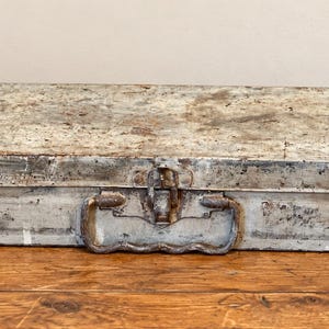 May include: A rectangular metal storage box with a weathered, distressed finish in shades of grey and white. The box features a central metal latch and a hinged handle on the front side, showing signs of oxidation and surface wear throughout.