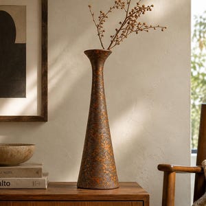 May include: A tall, slender, conical vase with a textured, mottled finish in shades of dark brown and metallic copper. The vase features a flared rim and a wide, stable base, containing a single dried branch with small, light brown buds.