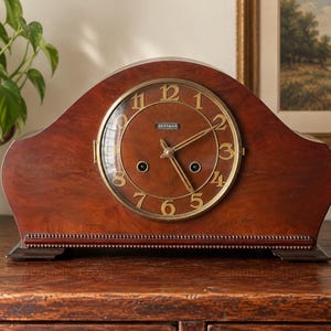 May include: A mantel clock with a polished mahogany-colored wooden case featuring a curved top. The circular face has a brass-toned bezel, gold-colored Arabic numerals, and two winding holes. The brand name "HERTMAR" is printed on the clock face.
