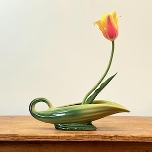 Könnte beinhalten: Eine grüne und gelbe Keramikvase mit einem gebogenen Griff und einer ausgestellten, bootsartigen Form. Eine einzelne Tulpe mit gelben und roten Blütenblättern und grünem Stiel und Blättern befindet sich in der Vase. Die Vase steht auf einer Holzoberfläche.
