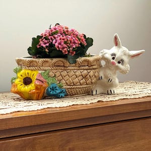 May include: A ceramic basket-shaped planter filled with pink flowers and green leaves. A white ceramic bunny figure sits beside the basket. A decorative ceramic sunflower and blue flowers are on the left. The items are on a wooden surface.