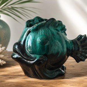 Könnte beinhalten: Eine keramische Fischvase in Türkis und Schwarz. Die Vase hat eine strukturierte, glänzende Oberfläche und eine breite Öffnung oben. Der Fisch hat detaillierte Flossen und ein rundes Auge. Die Vase ist ein dekorativer Gegenstand.